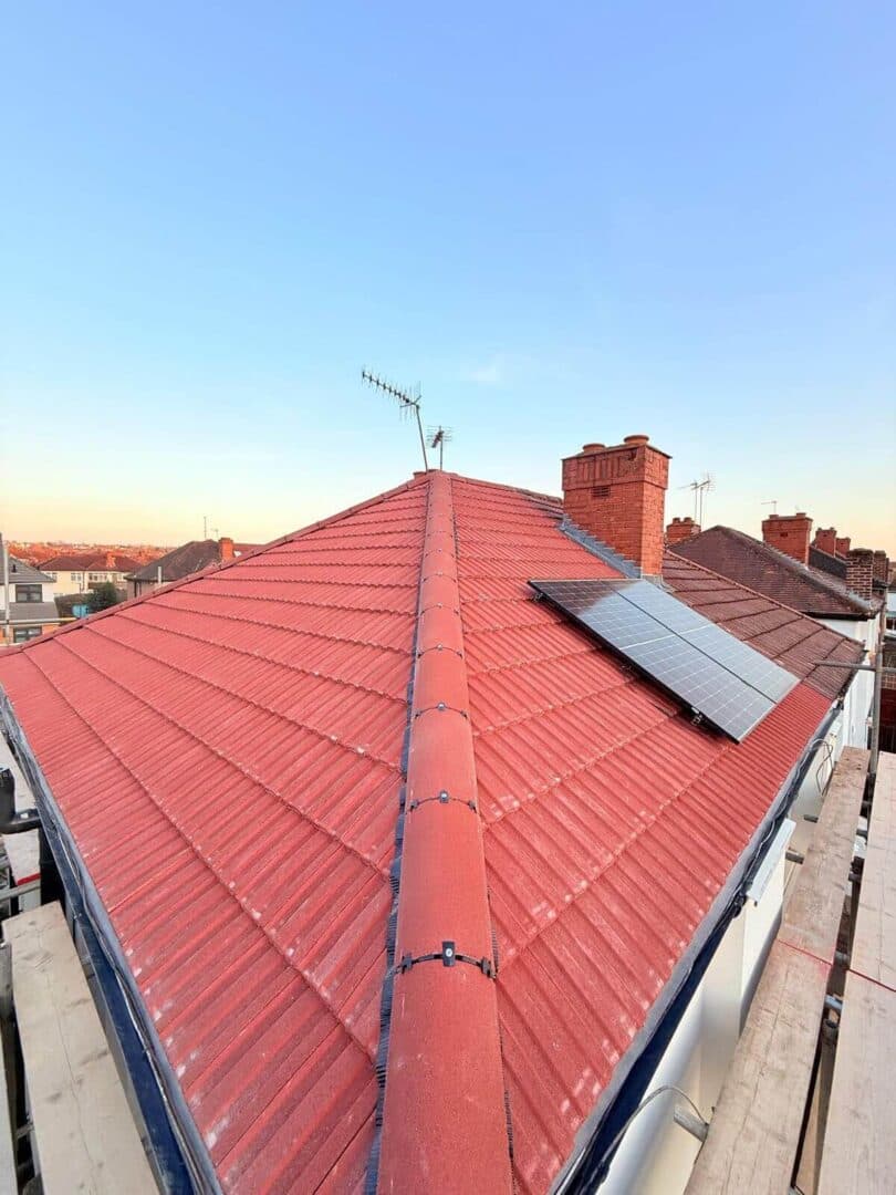 A red concrete-tiled hip roof with a dry-fix ridge and solar panels on one slope, photographed from the scaffold
